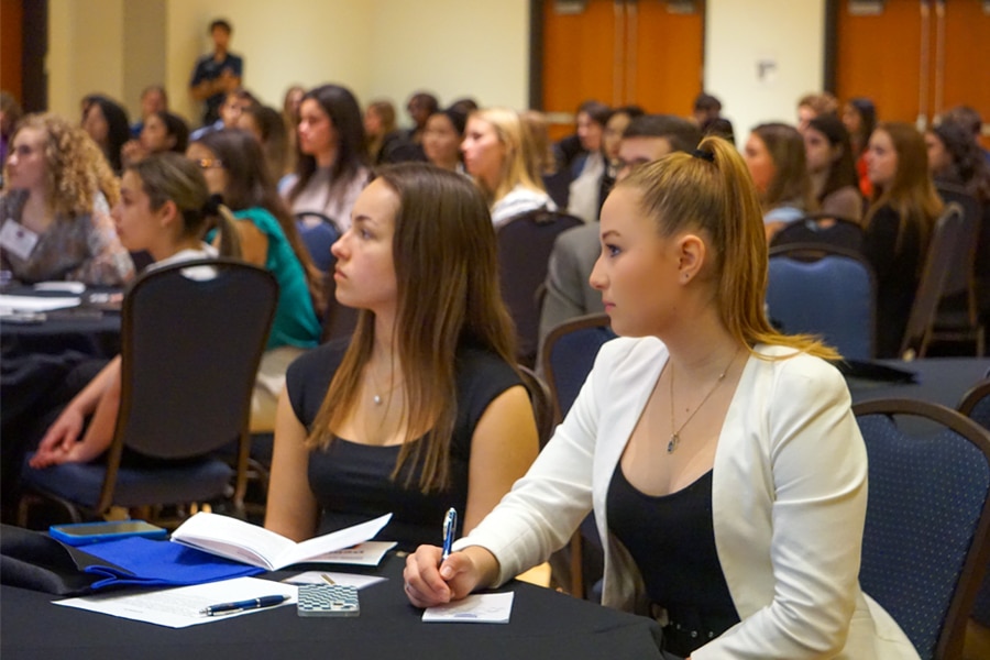 Students seated at round tables attentively listening, with notebooks and pens in hand for note-taking during a professional development event.
