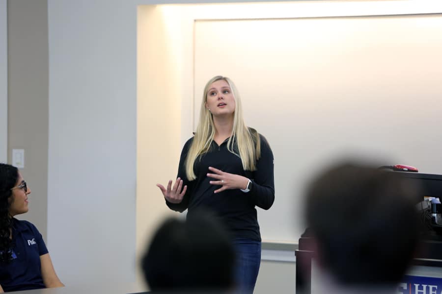 A professional woman with long blonde hair stands confidently at the front of a classroom speaking to an audience, while a seated woman listens attentively.