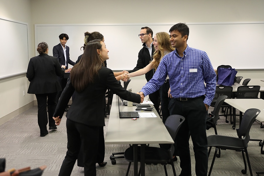 Students and business professionals shake hands across a long table in a modern conference room as part of a collaborative meeting.