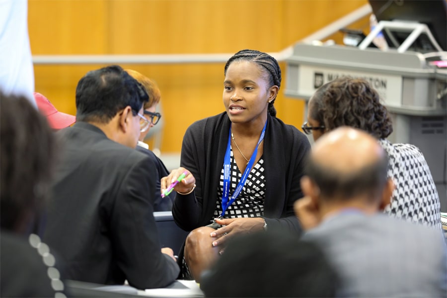 A woman in a black-and-white dress with a blue lanyard speaks to a small group in a collaborative setting, sharing her insights with colleagues during a research workshop.