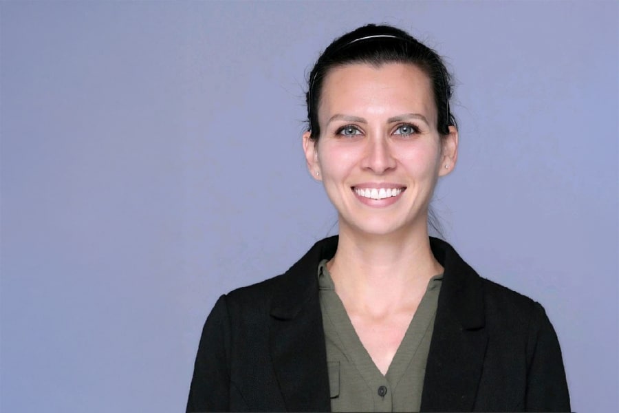 Portrait of Rachael Kistner smiling, wearing a black blazer and olive green blouse, with her hair pulled back against a plain gray background.