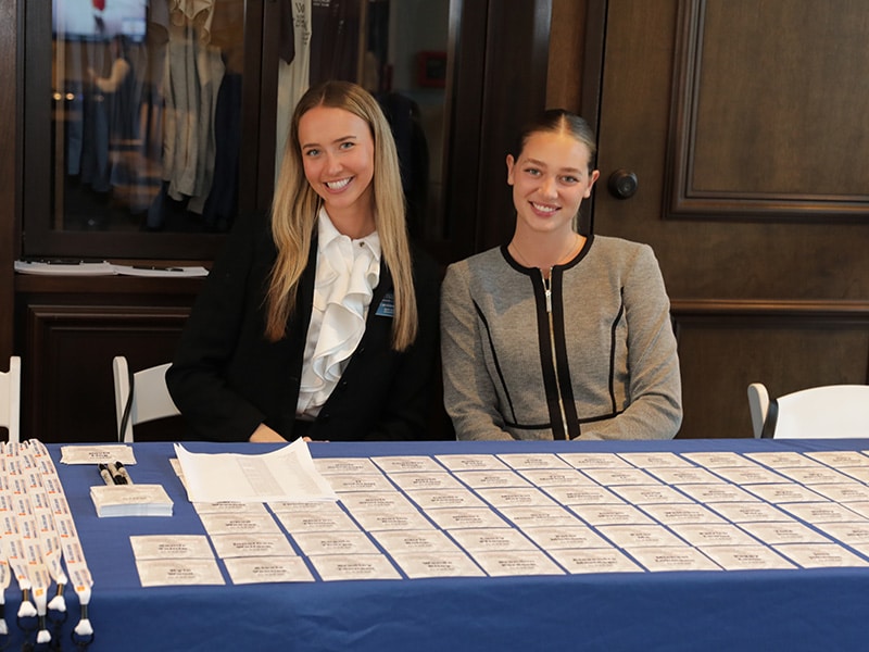 Two young women smiling at the camera from behind a conference welcome table filled with name tags.