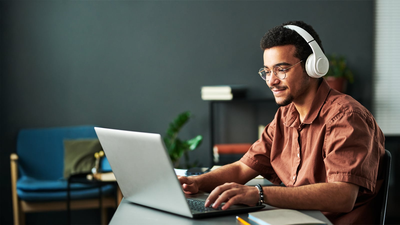 A young man wearing glasses and white headphones types on a laptop in a bright, modern study space with a blue sofa and plants in the background.