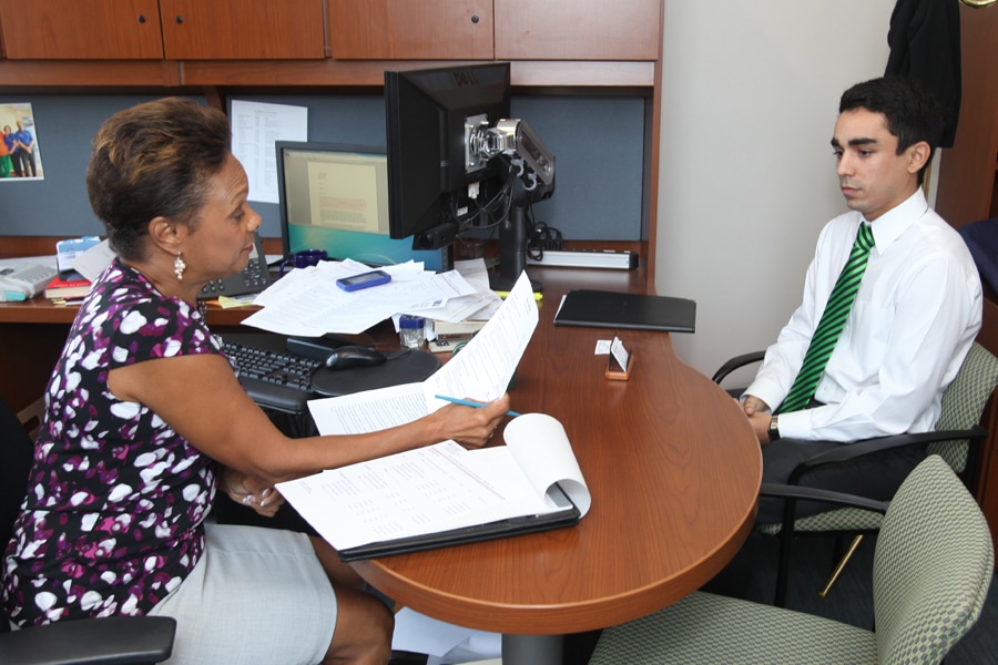 An academic advisor reviews documents while speaking with a formally dressed student sitting across a desk in an office.