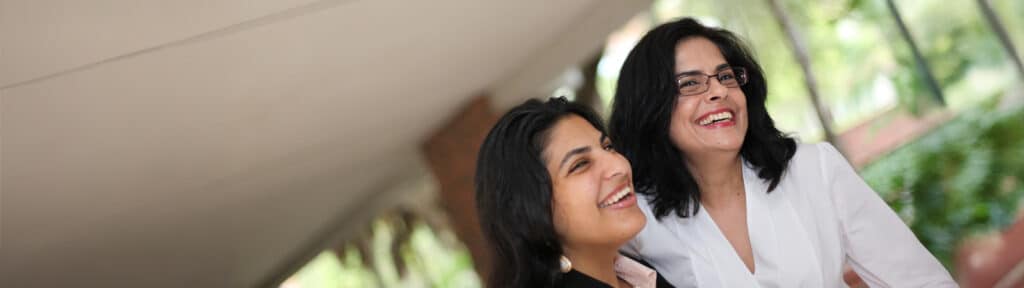 Two professional women stand smiling together outdoors in a shaded area at the Warrington College of Business, with greenery and a red brick building in the background.