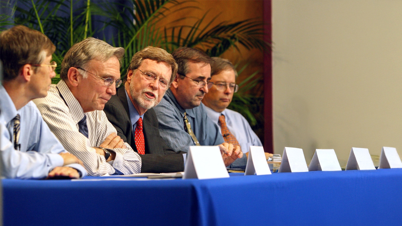 A group of distinguished men engaged in conversation during a panel discussion seated at a long table with blue tablecloth and blank name cards.