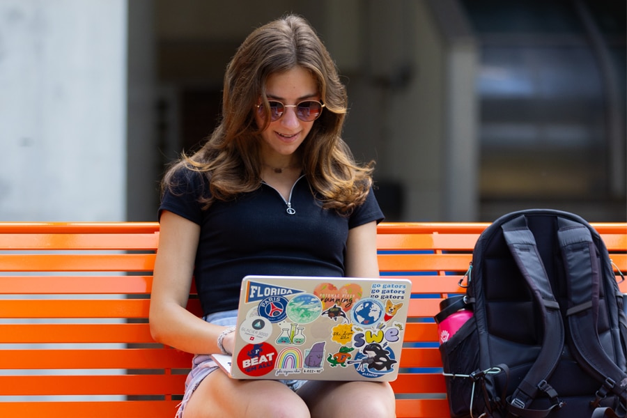 A student wearing sunglasses sits on an orange bench outdoors, smiling as she reviews her sticker-covered laptop. A backpack sits beside her.