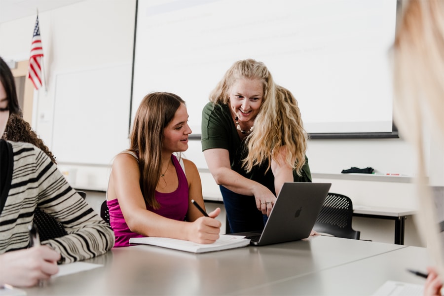 A faculty advisor leans over a seated student, pointing at a laptop screen while the student smiles and takes notes in a classroom setting.