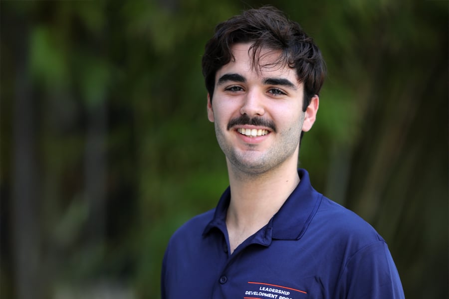 A young man with dark hair and a mustache smiles at the camera, wearing a navy blue Leadership Development Program polo shirt, with greenery in the background.