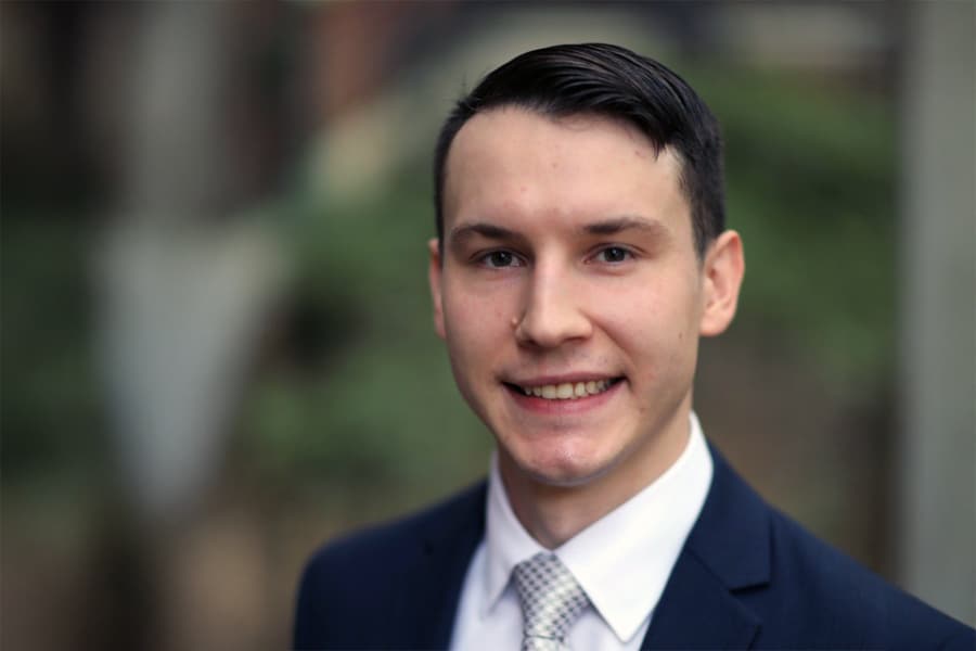 Portrait of Sam Bodman, a young man wearing a navy suit, white shirt, and gray patterned tie, smiling with a blurred natural background.