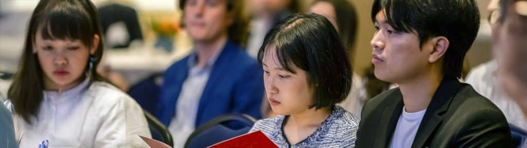 A diverse group of focused University of Florida students attentively listens during a presentation, some taking notes and reviewing materials.