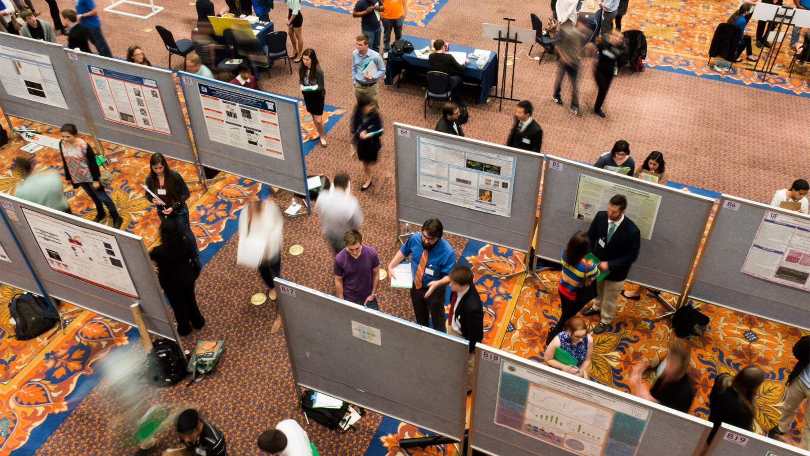 An overhead view of students and faculty walking and standing among rows of research posters at an academic symposium held in a large, carpeted room with a floral pattern.