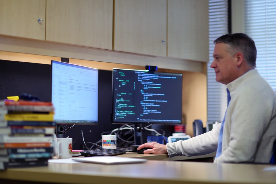 Warrington College of Business faculty member seated at a desk working on a computer with two monitors displaying email and coding content. A stack of business books is on the desk nearby.
