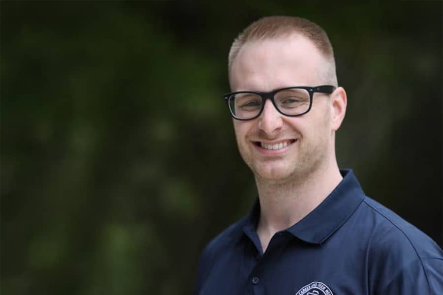 Portrait of Seth Hampsey, a young man wearing glasses and a navy blue collared shirt, smiling outdoors against a blurred natural background.