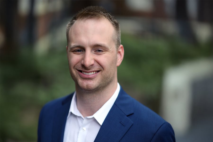 Portrait of Seth Hampsey, a young man wearing a blue suit jacket and white shirt, smiling outdoors with a blurred green background.