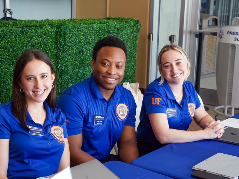 Three smiling student ambassadors wearing blue shirts with the Warrington College of Business logo sit behind a table covered with a blue tablecloth, ready to help visitors.