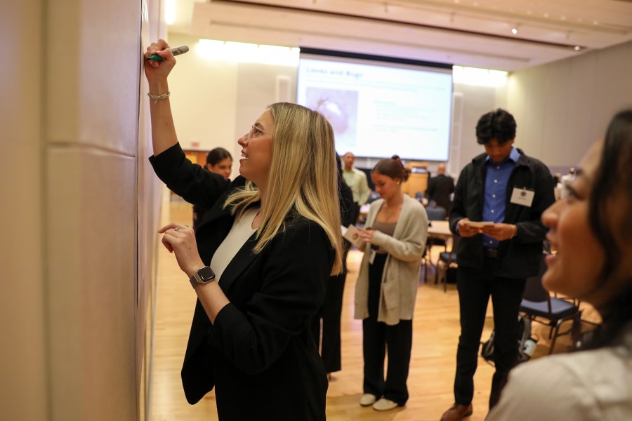A young woman in business attire writes on a whiteboard with a marker while others watch and interact in the background during a professional workshop.