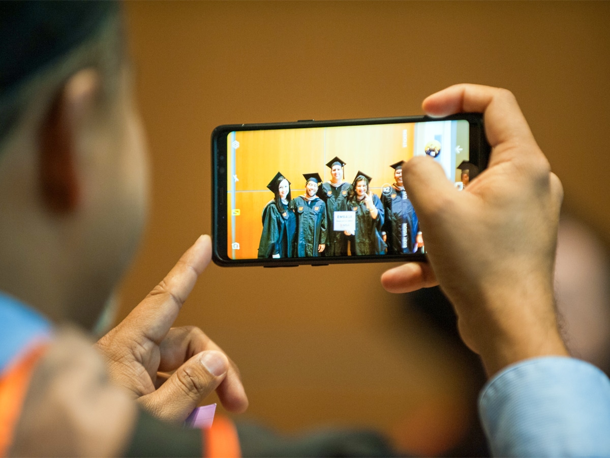 A graduate in cap and gown holds up a smartphone to photograph a group of fellow graduates standing against a wooden-paneled wall.