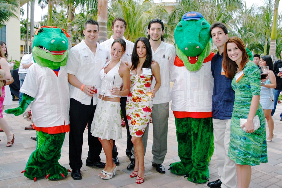 A group of young adults posing outdoors with two Gator mascots, all smiling and dressed in casual to semi-formal attire, surrounded by palm trees.