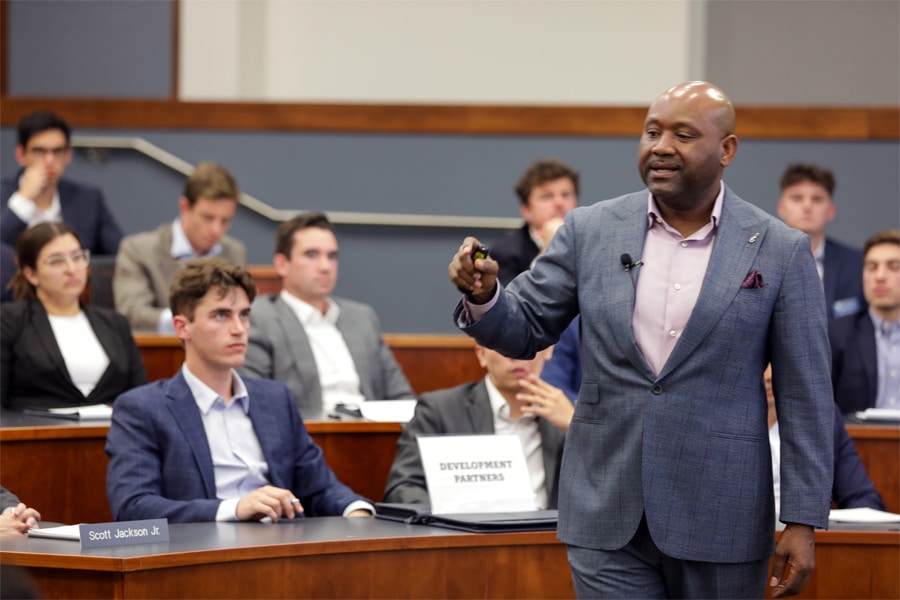 A well-dressed speaker in a gray suit presents to an attentive audience of graduate students seated in a lecture hall.