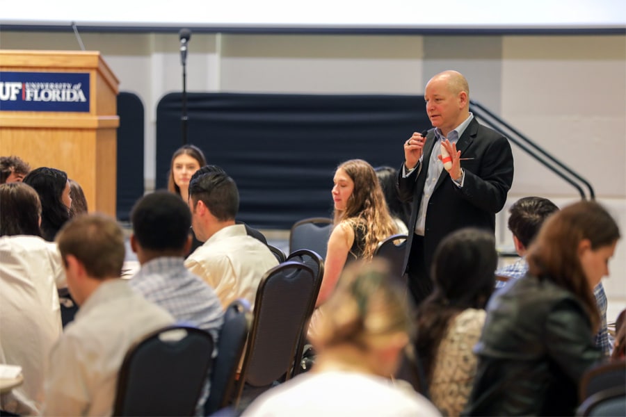 A speaker in a dark suit holds a microphone and addresses a seated audience of students at a Warrington Welcome event in a University of Florida venue.