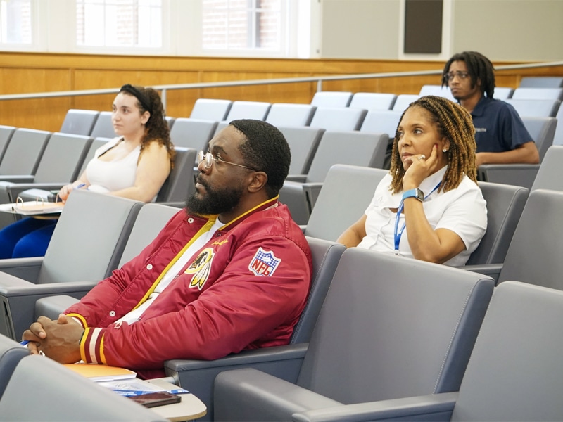 Several individuals seated in a lecture hall, listening attentively, including a man in a red NFL jacket and a woman in a white shirt resting her chin on her hand.