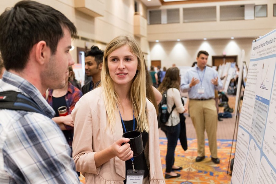A young woman holding a black mug discusses her research with a fellow attendee near a large poster presentation at a research symposium.