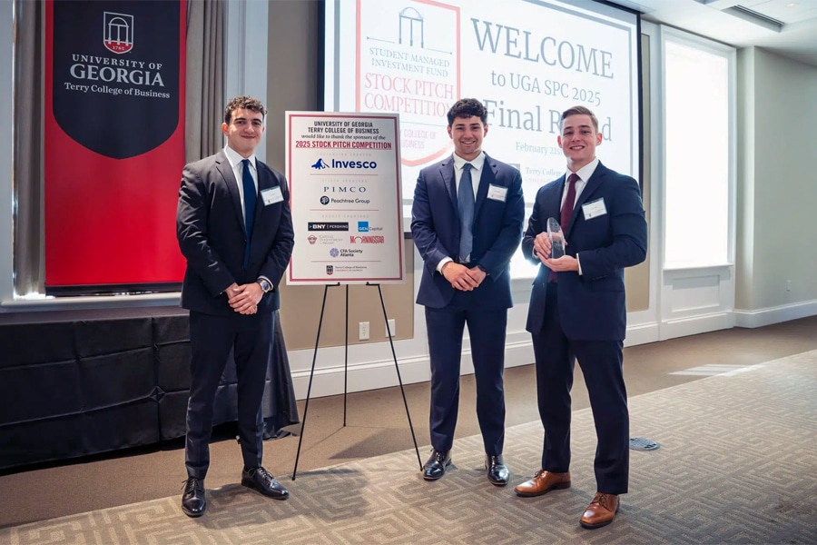 Three graduate finance students in business attire stand together at the University of Georgia’s Stock Pitch Competition, one holding a trophy, with event signage and a presentation screen behind them.