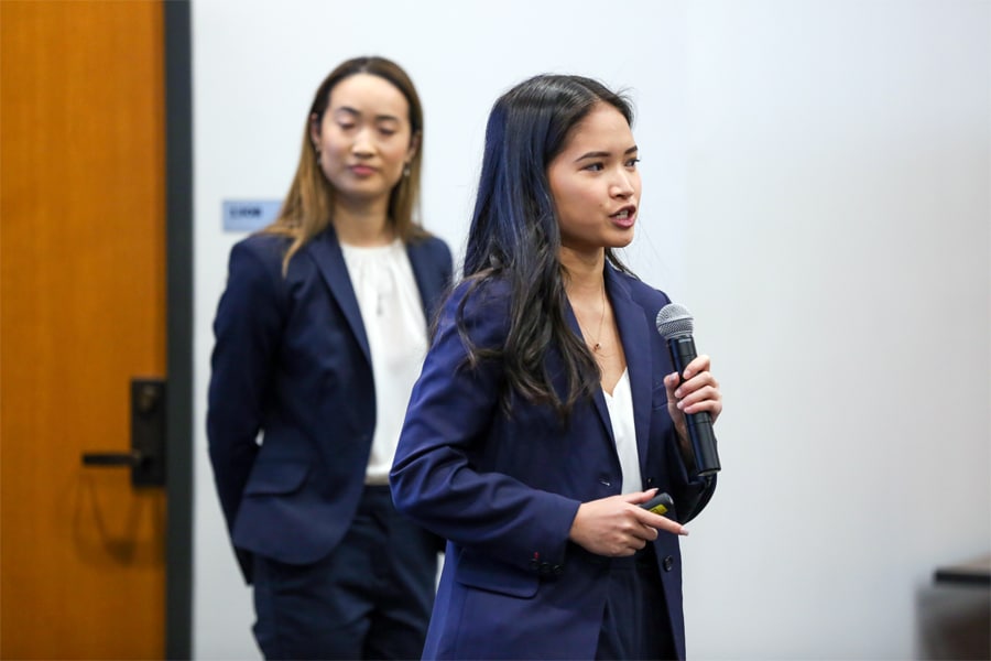 A business student in a suit presents with a microphone while another student stands attentively in the background.
