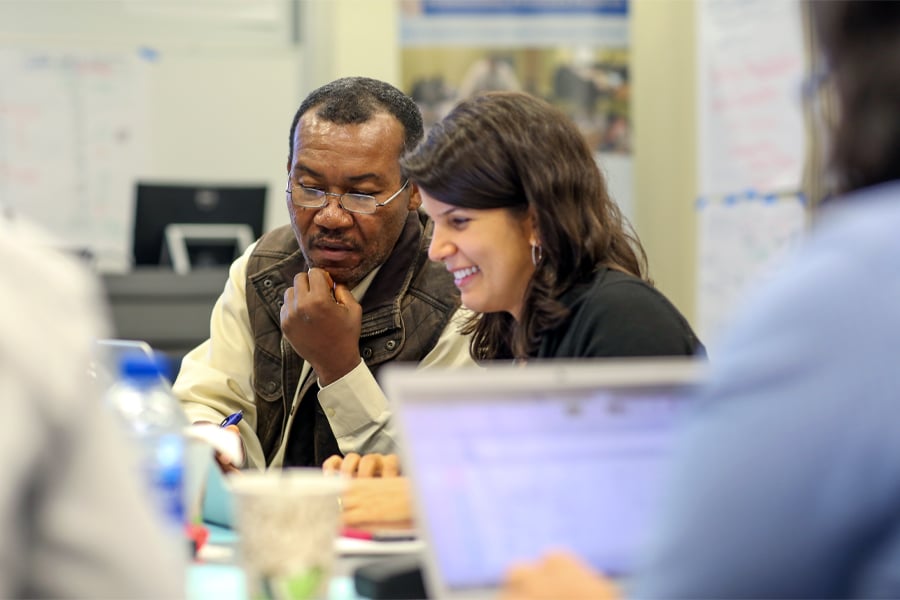 A man and a woman collaborate on a project in a classroom setting. They are engaged and focused, with notebooks and laptops open on the table.