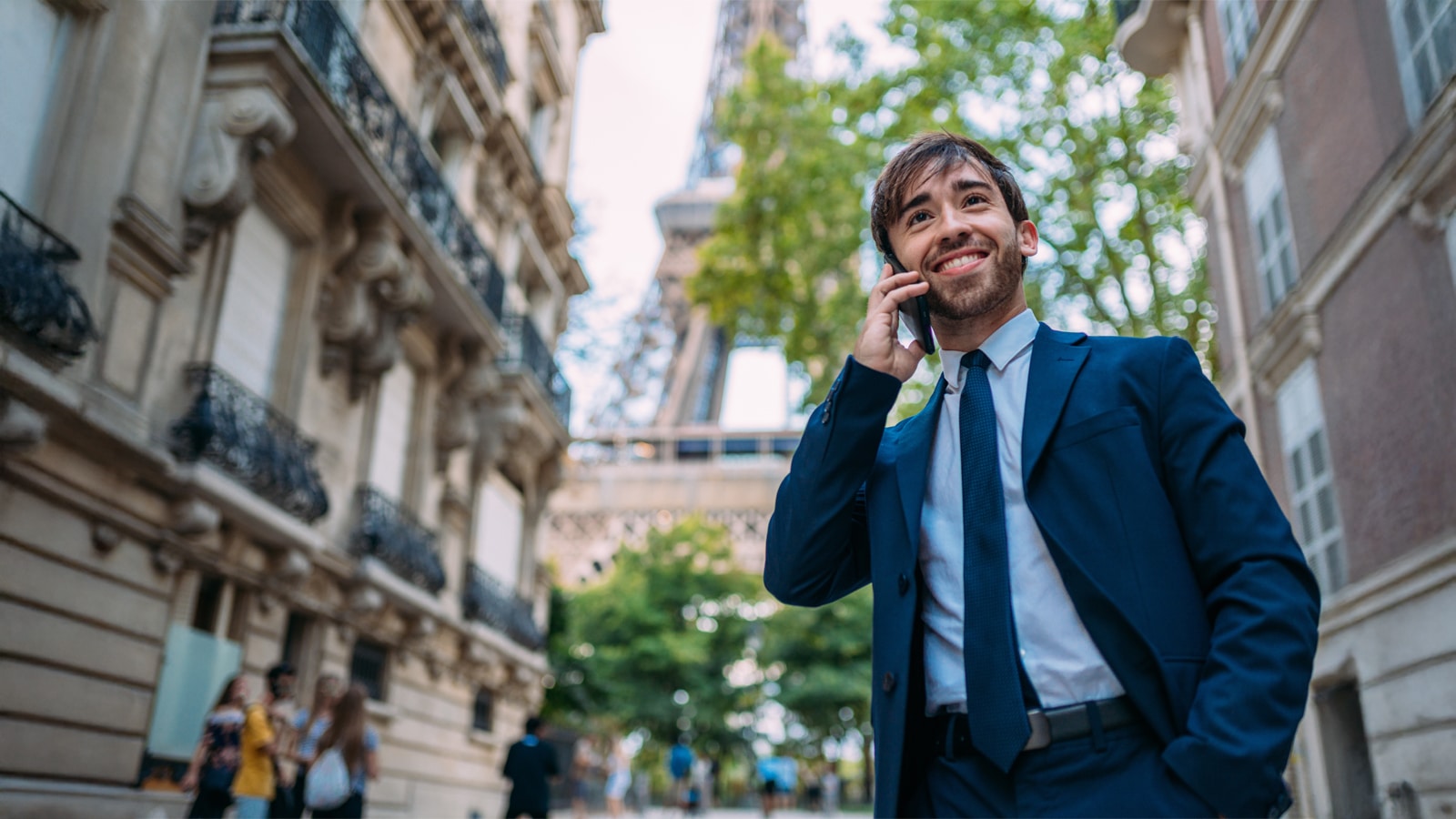 A smiling young man in a blue suit and tie talks on his phone on a Paris street, with the Eiffel Tower visible in the background framed by buildings and trees.