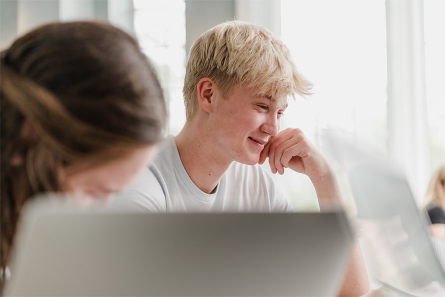 A young student with blonde hair smiles while looking at a laptop screen in a bright room.