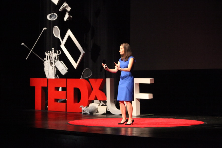 A woman in a blue dress speaks on stage at TEDxUF, standing on the iconic red circle with large TEDxUF letters behind her.