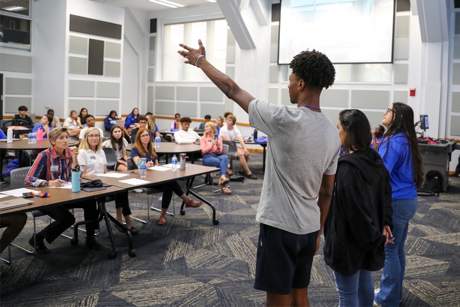 A young man stands at the front of a classroom with two peers beside him, speaking to an attentive group of seated adults and students.