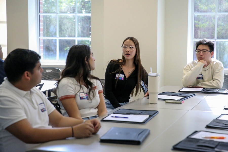 Four students sit around a table in a bright classroom, actively discussing ideas. One student, wearing glasses, speaks while others listen and take notes.