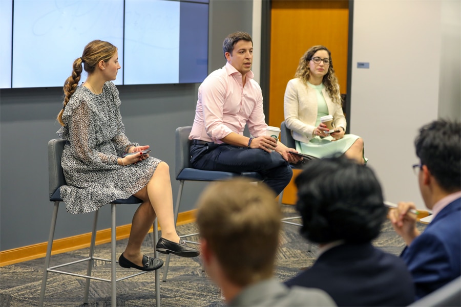 Three young professionals sit on stools at the front of a classroom, leading a discussion with a group of students who listen attentively.