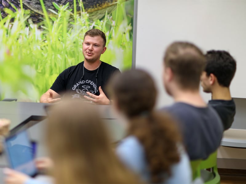 A young man in a black t-shirt is speaking animatedly to a group of students seated around a table in a bright room with a large, nature-themed mural on the wall.