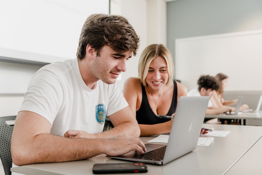 Two college students, a young man and a young woman, sit side-by-side at a classroom table, collaborating on a laptop as they discuss their research topic.