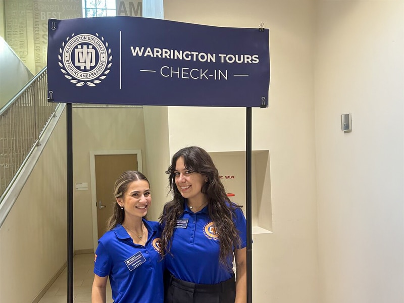 Two Warrington Diplomats, wearing matching blue shirts, smile and pose under a “Warrington Tours Check-In” sign inside a building at the University of Florida.