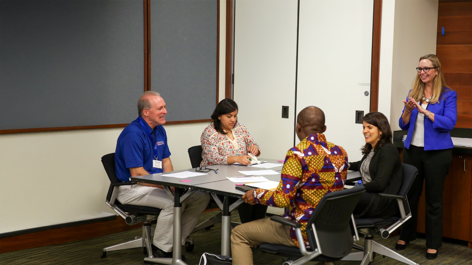 A group of professionals sit around a conference table, engaged in a lively discussion, with a facilitator in a blue jacket standing and applauding.
