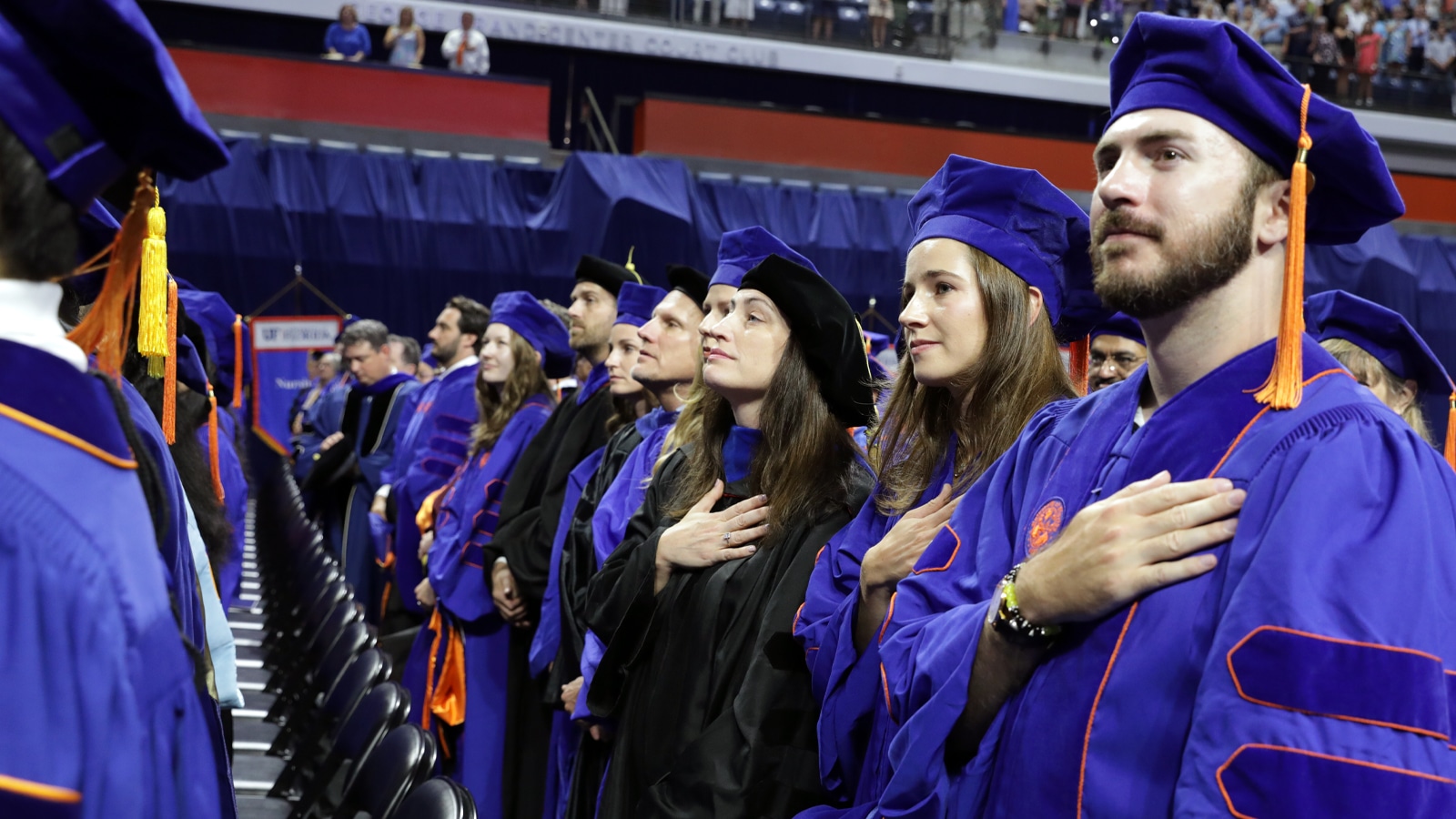 Doctoral graduates in blue and orange regalia stand with hands over their hearts during a University of Florida commencement ceremony.