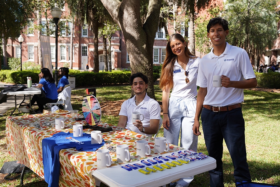 Students from the Warrington College of Business' Business College Council stand near a table with mugs and snacks at an outdoor event