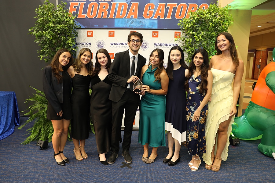 Wearing dress clothes, undergraduate students from the Warrington College of Business pose for a picture in front of a wall with the University of Florida school logo