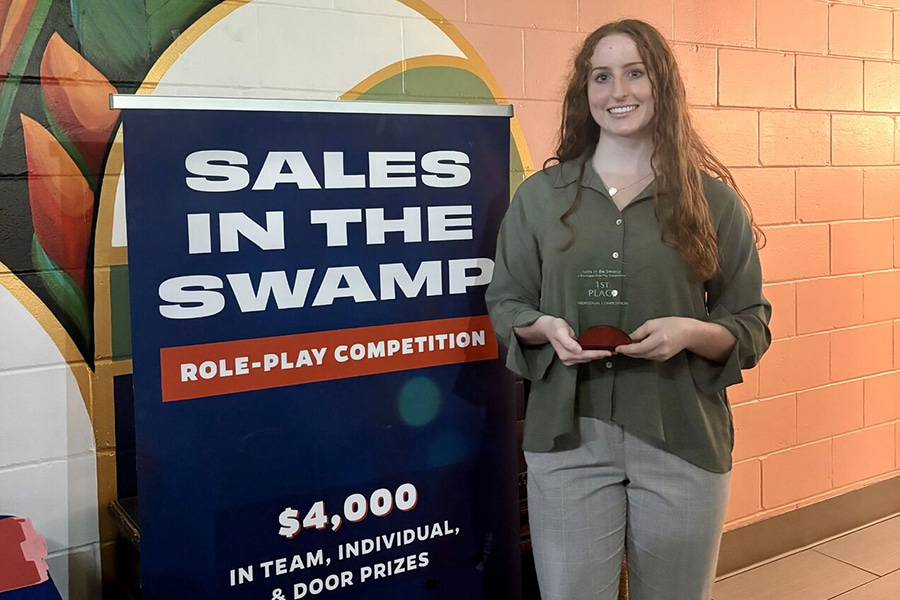 A student holds their award while standing next to a poster for Sales in the Swamp, a competition put on by the UF Warrington College of Business
