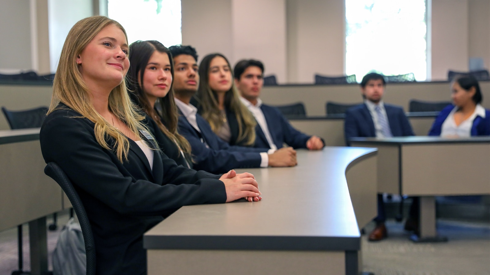 Group of undergraduate students attentively listening in a tiered classroom, dressed in professional attire.