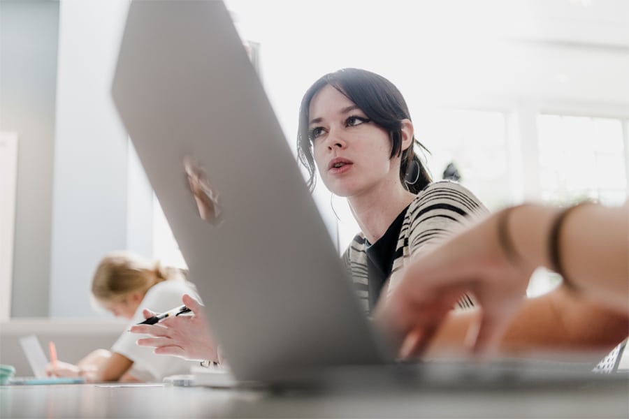 A focused young woman with dark hair and winged eyeliner gestures while speaking in a brightly lit classroom, a laptop screen partially obscuring her face.