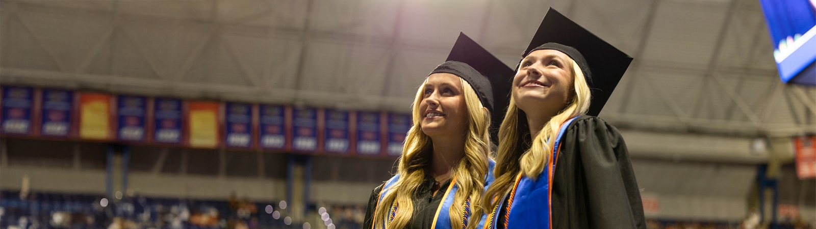 Two female graduates wearing caps and gowns with blue and orange honor cords look upward with smiles inside a large stadium filled with banners.