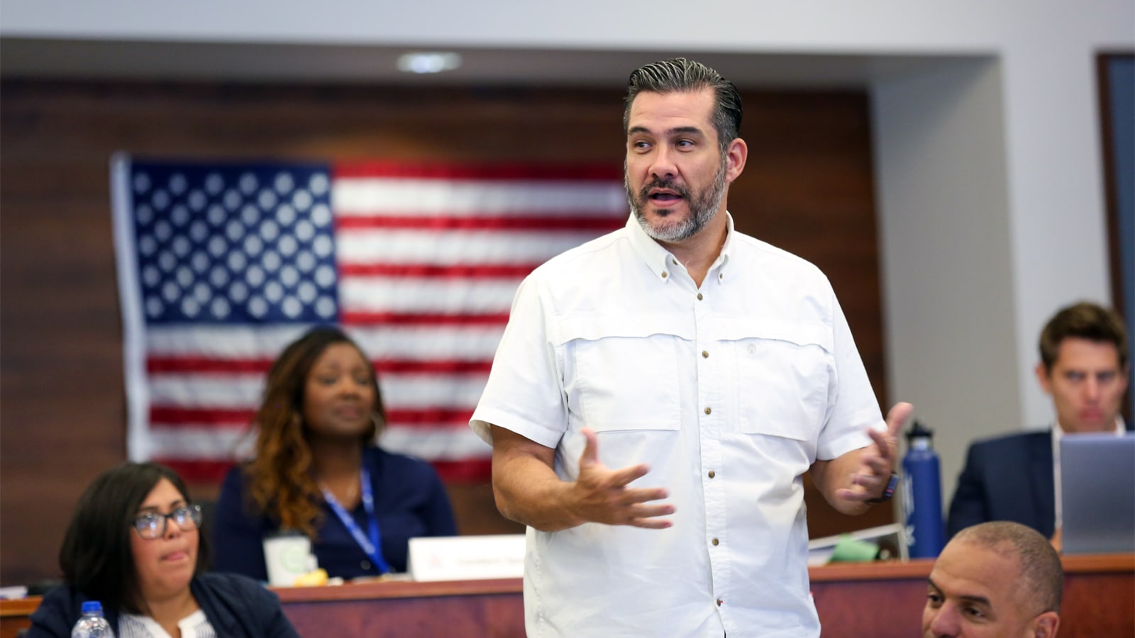 A man in a white short-sleeve shirt speaks confidently in front of a classroom with a blurred American flag in the background. Several people listen attentively in the background.