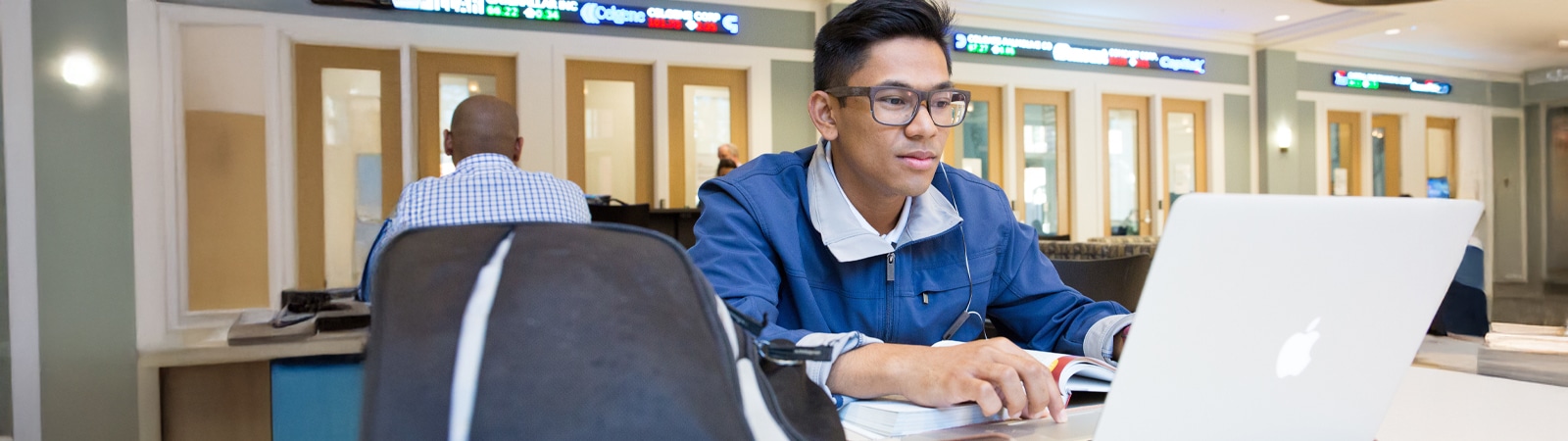 A focused student studies at a laptop in a business school setting, with stock market tickers displayed on screens behind.