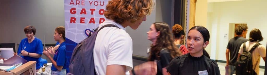 Two Warrington students engage in conversation near a tabling event with blue-shirted volunteers and other students milling about in the background.